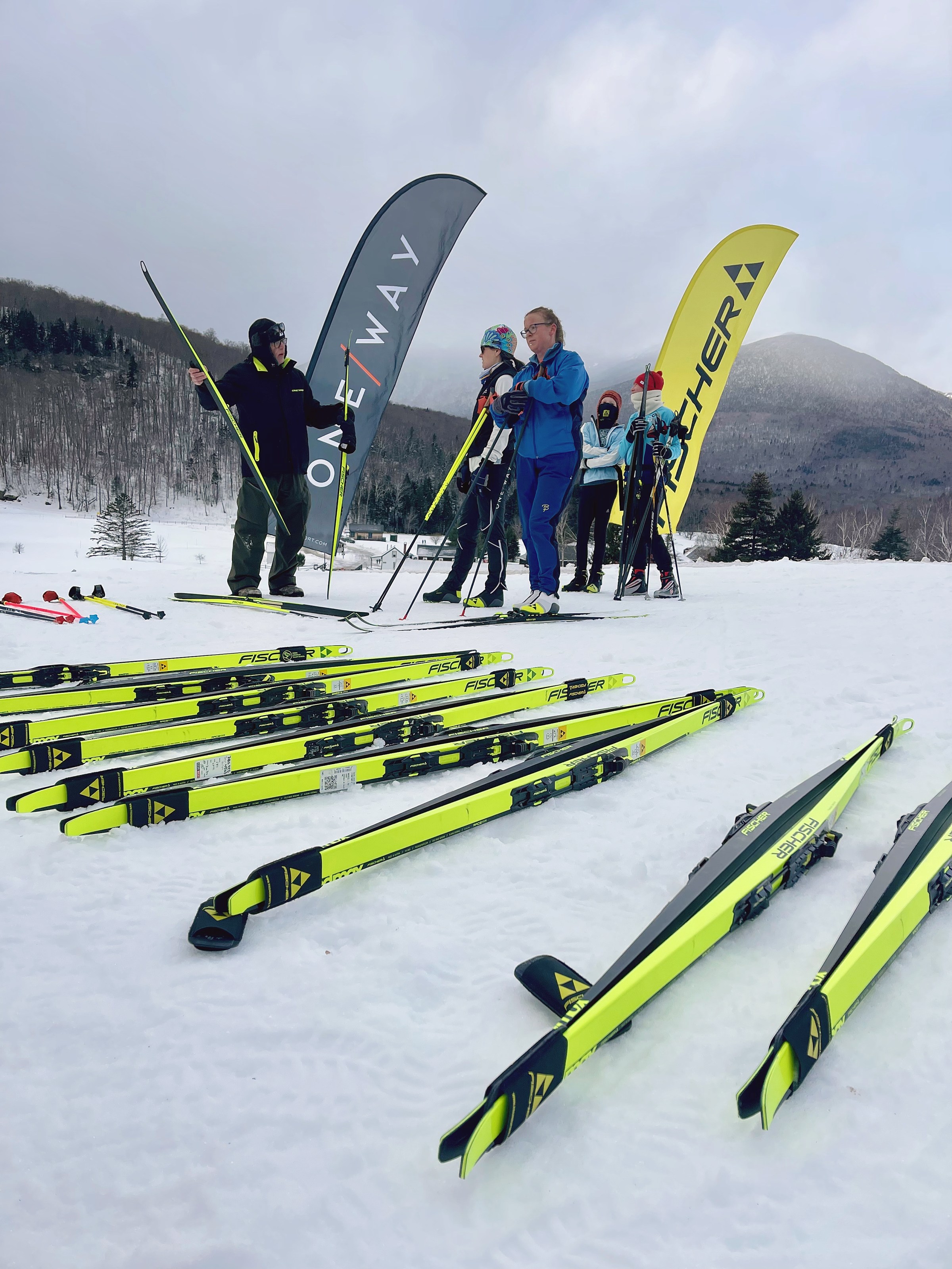 People in winter gear stand with skis and poles on snowy ground, with mountains and banners in the background.