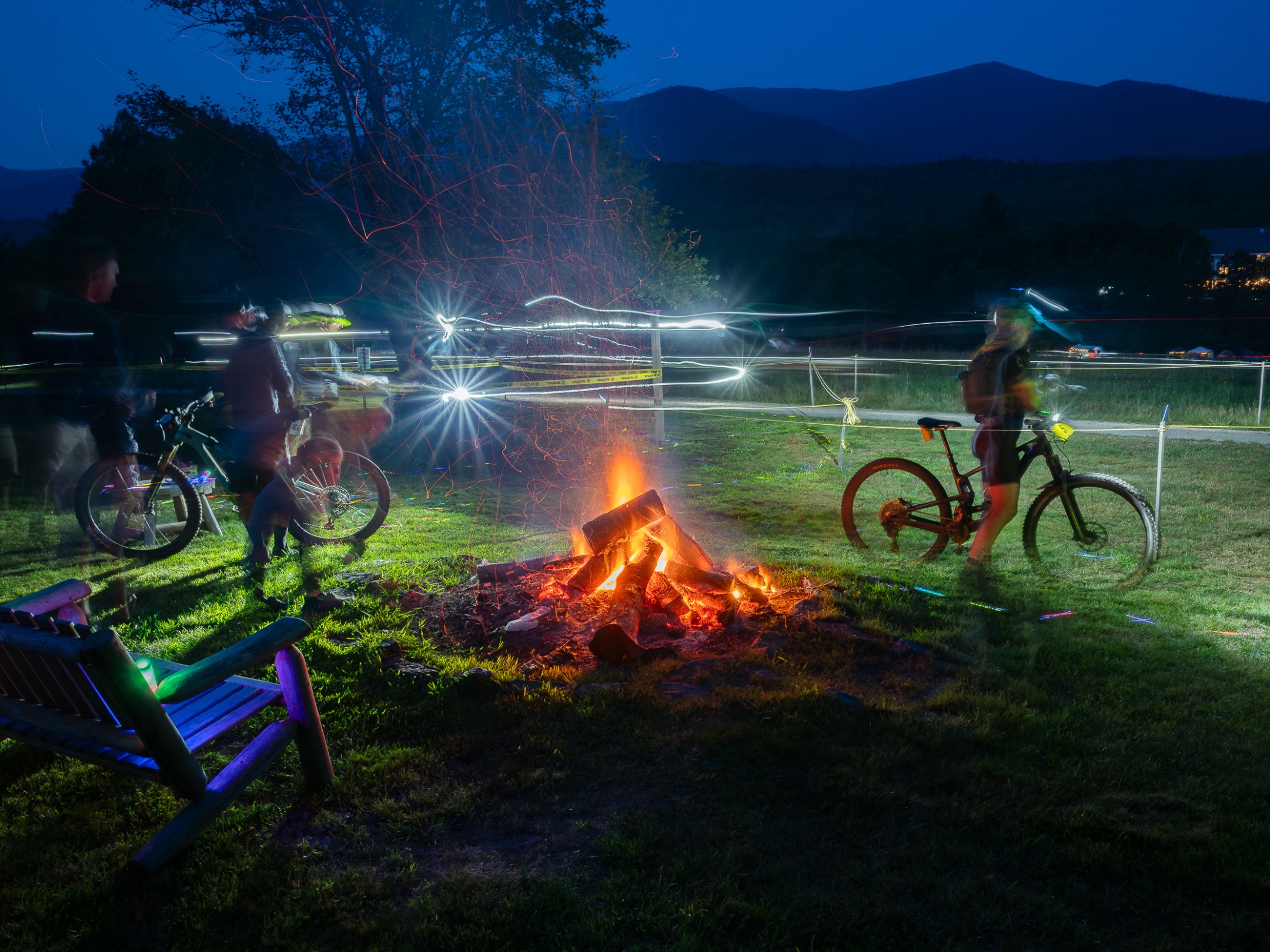 Night scene with bikes and people around a campfire, light trails and mountains in the background.