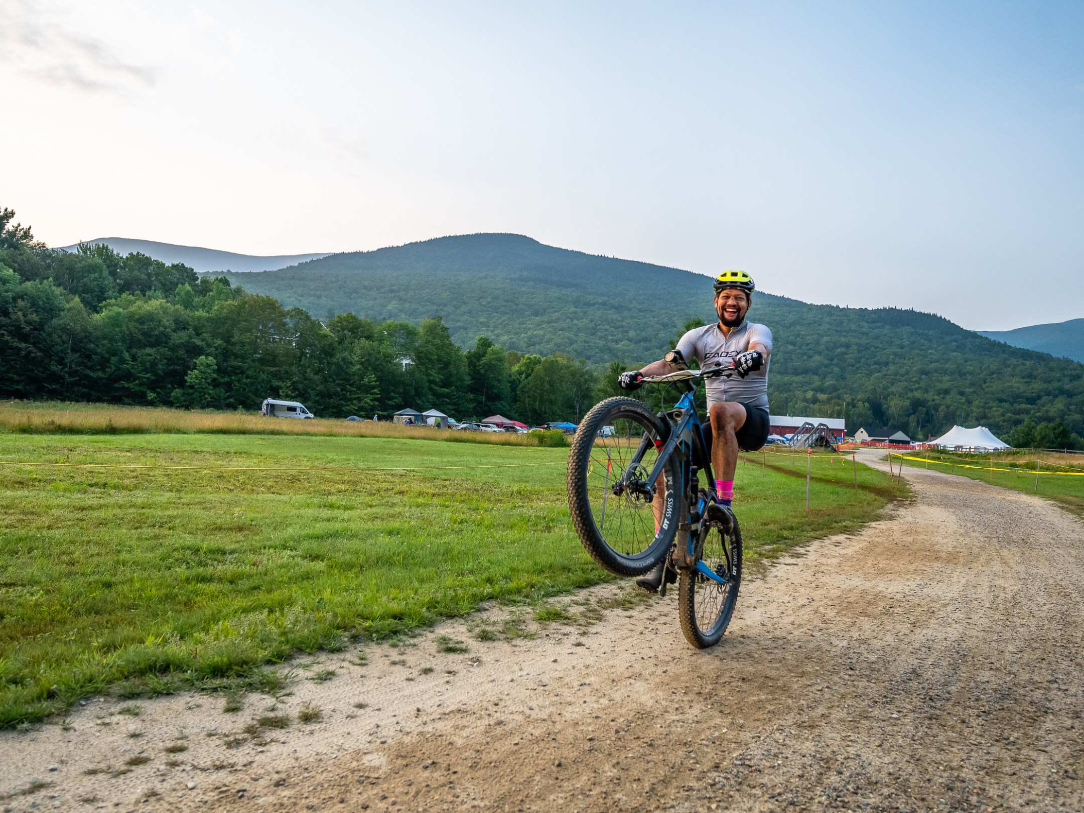 Cyclist performing a wheelie on a dirt path with mountains and tents in the background.