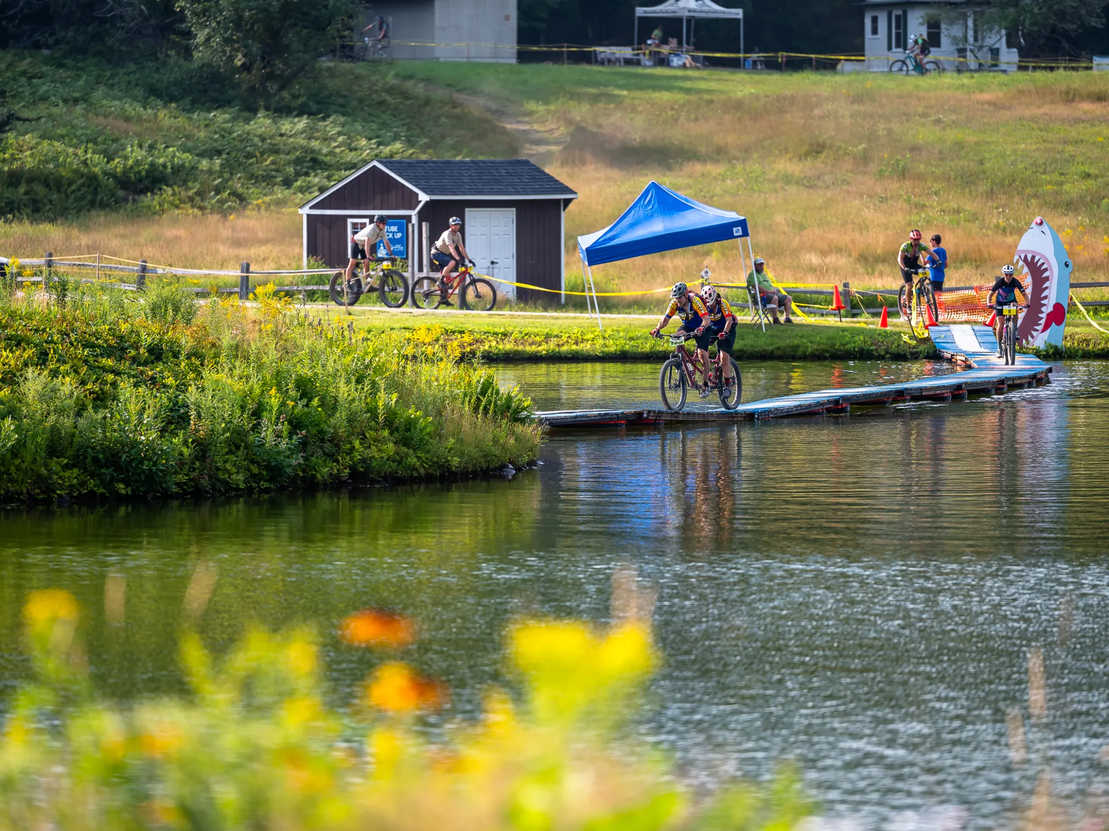 Cyclists cross a floating bridge on a pond during an outdoor event.