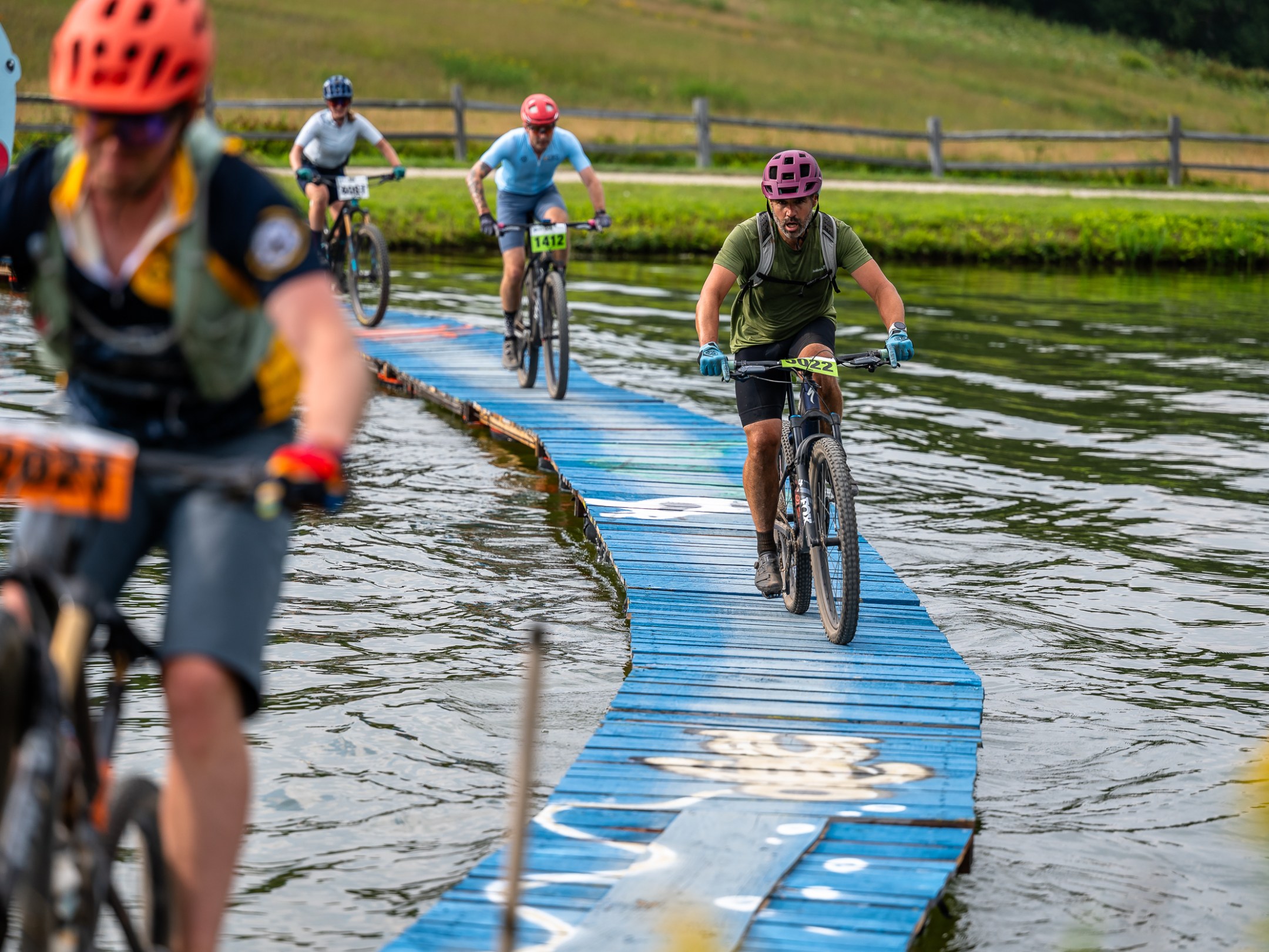 Cyclists on a narrow blue bridge over water in a race, wearing helmets and numbered bibs.