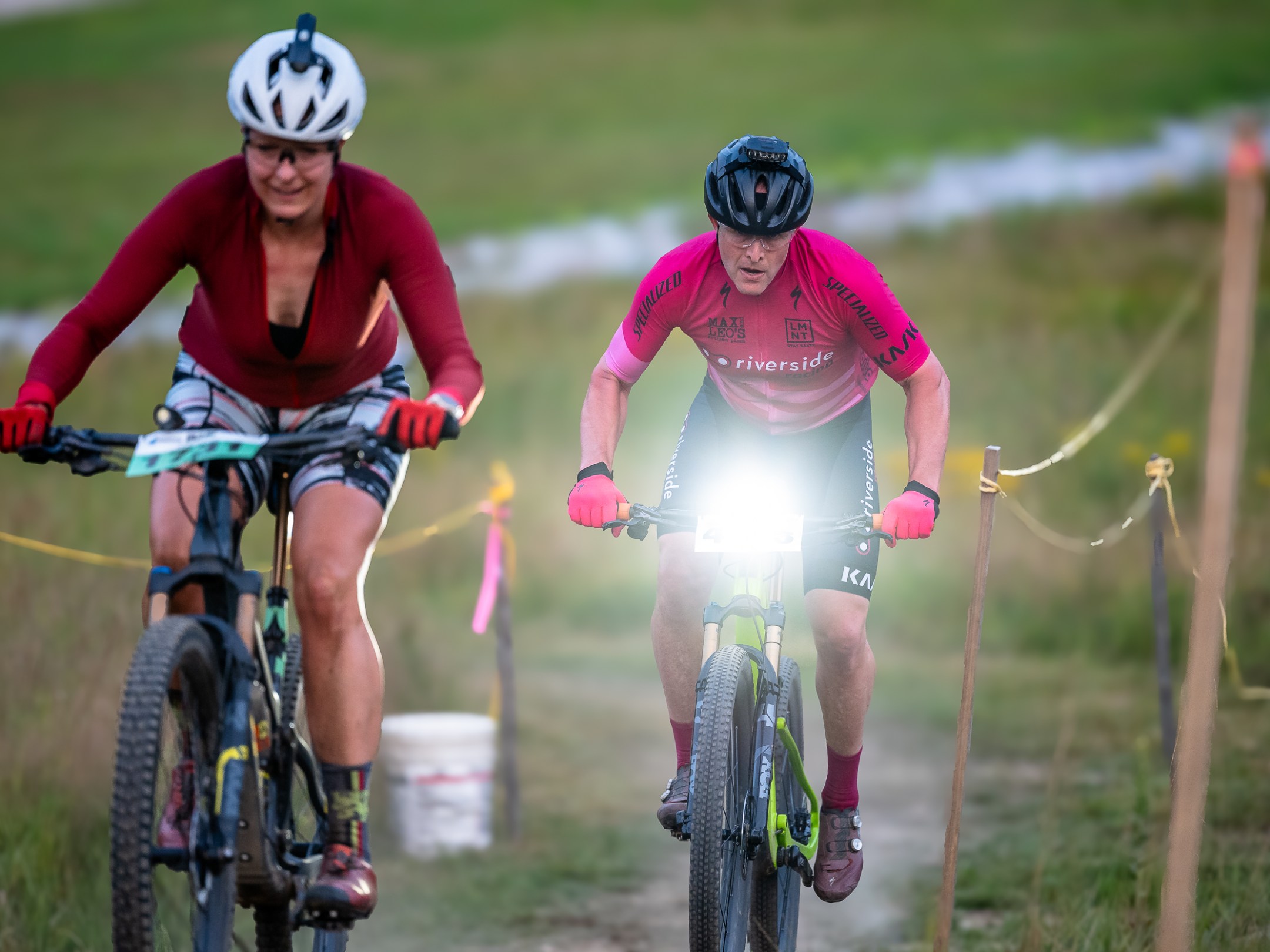 Two cyclists riding mountain bikes on a trail with lush green grass.