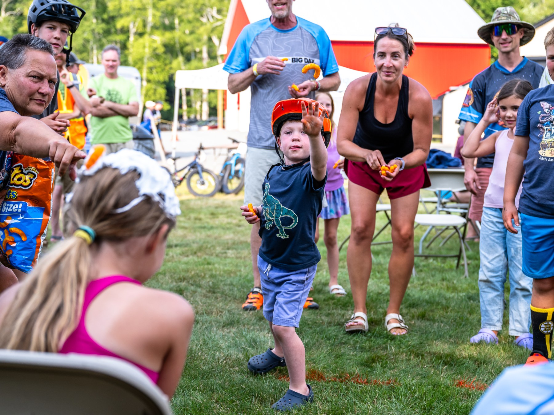 Child in helmet throwing object at girl with whipped cream on her head, surrounded by onlookers in an outdoor setting.