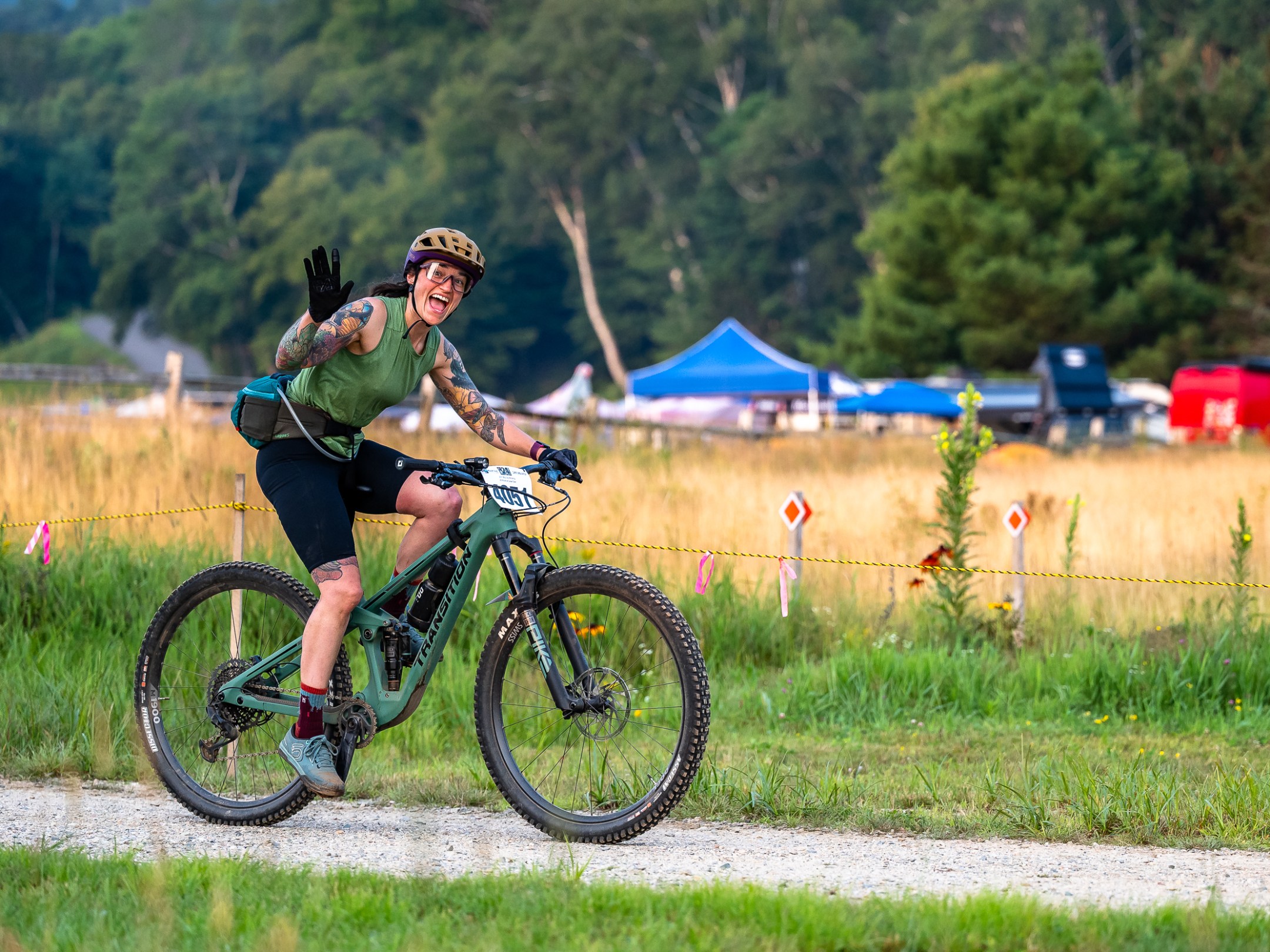 Cyclist waving on a trail with tents and trees in the background.