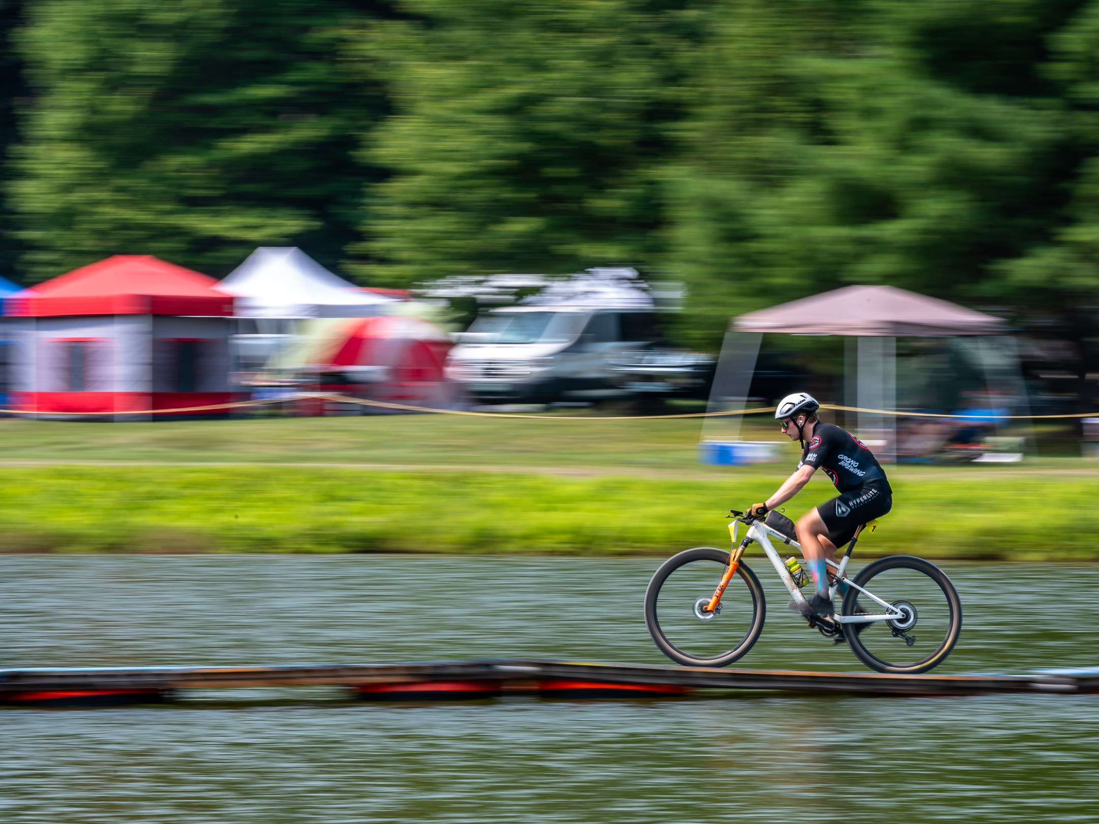 Person riding a bicycle on a narrow path over water with tents in the background.
