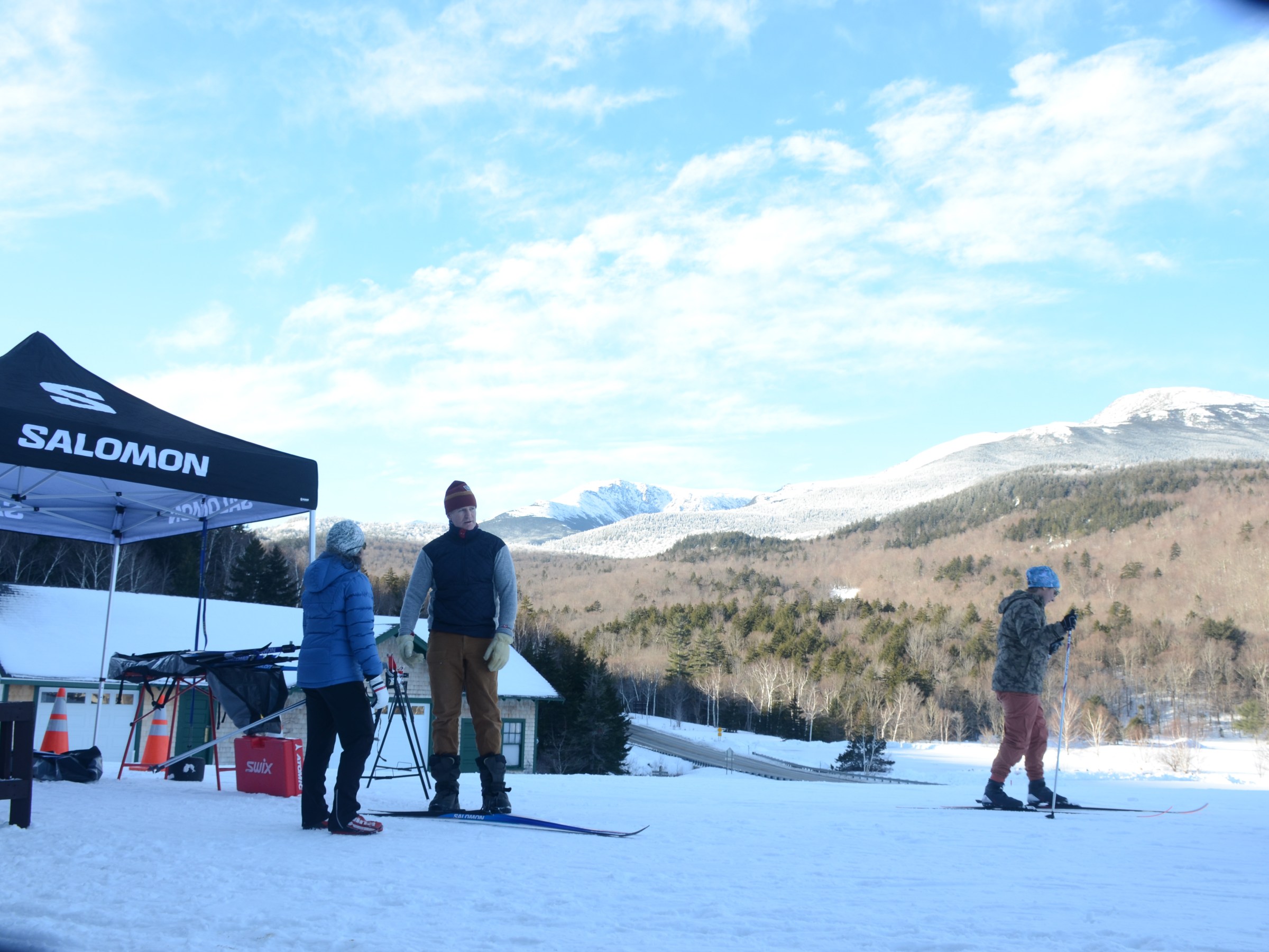 People in winter gear stand and ski near a Salomon tent on a snowy mountain.
