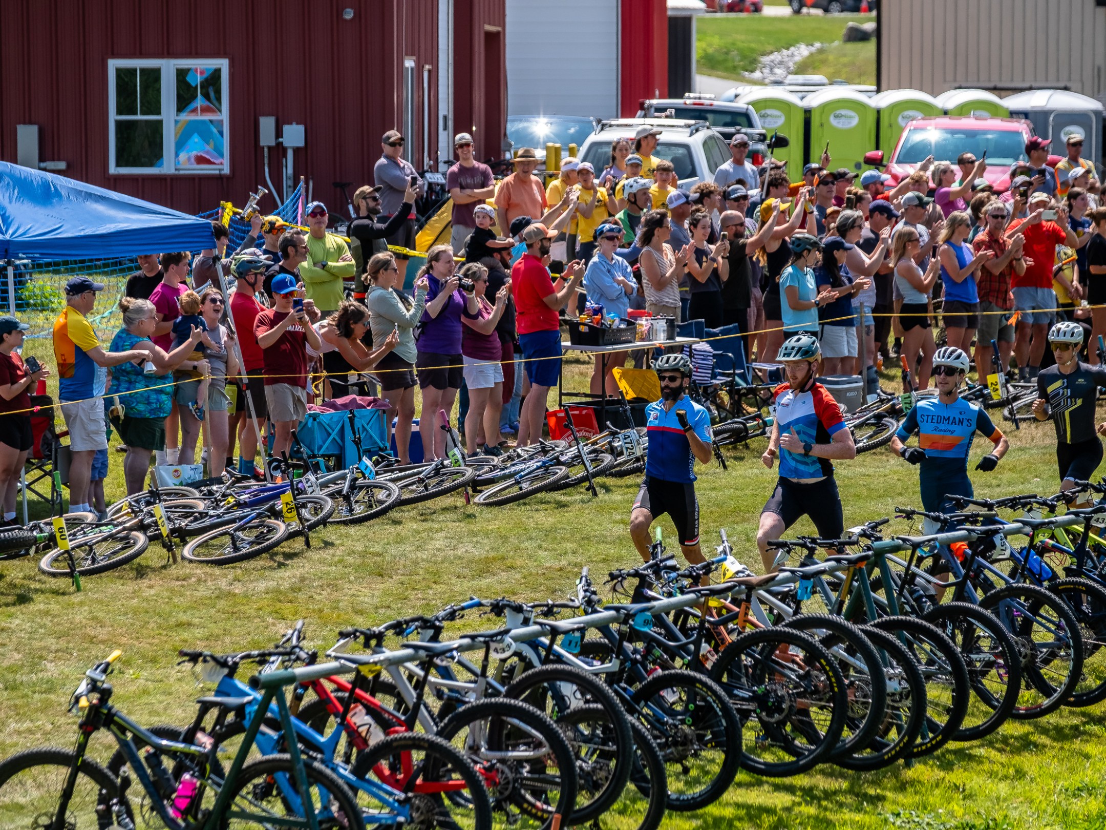 Cyclists run beside lined-up mountain bikes; crowd cheers from behind a barrier at an outdoor event.