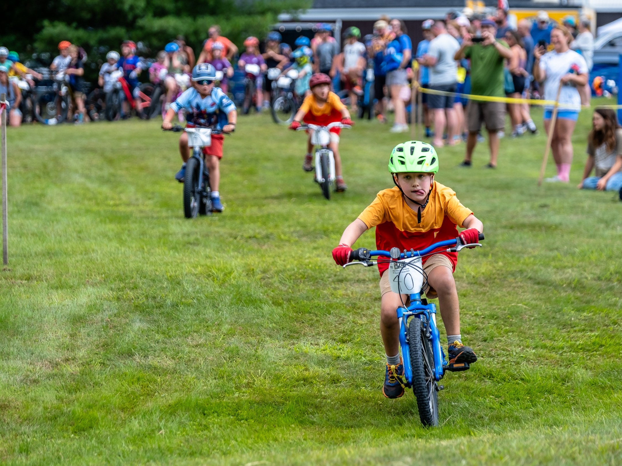 Kids racing on bikes in a grassy area with spectators in the background.