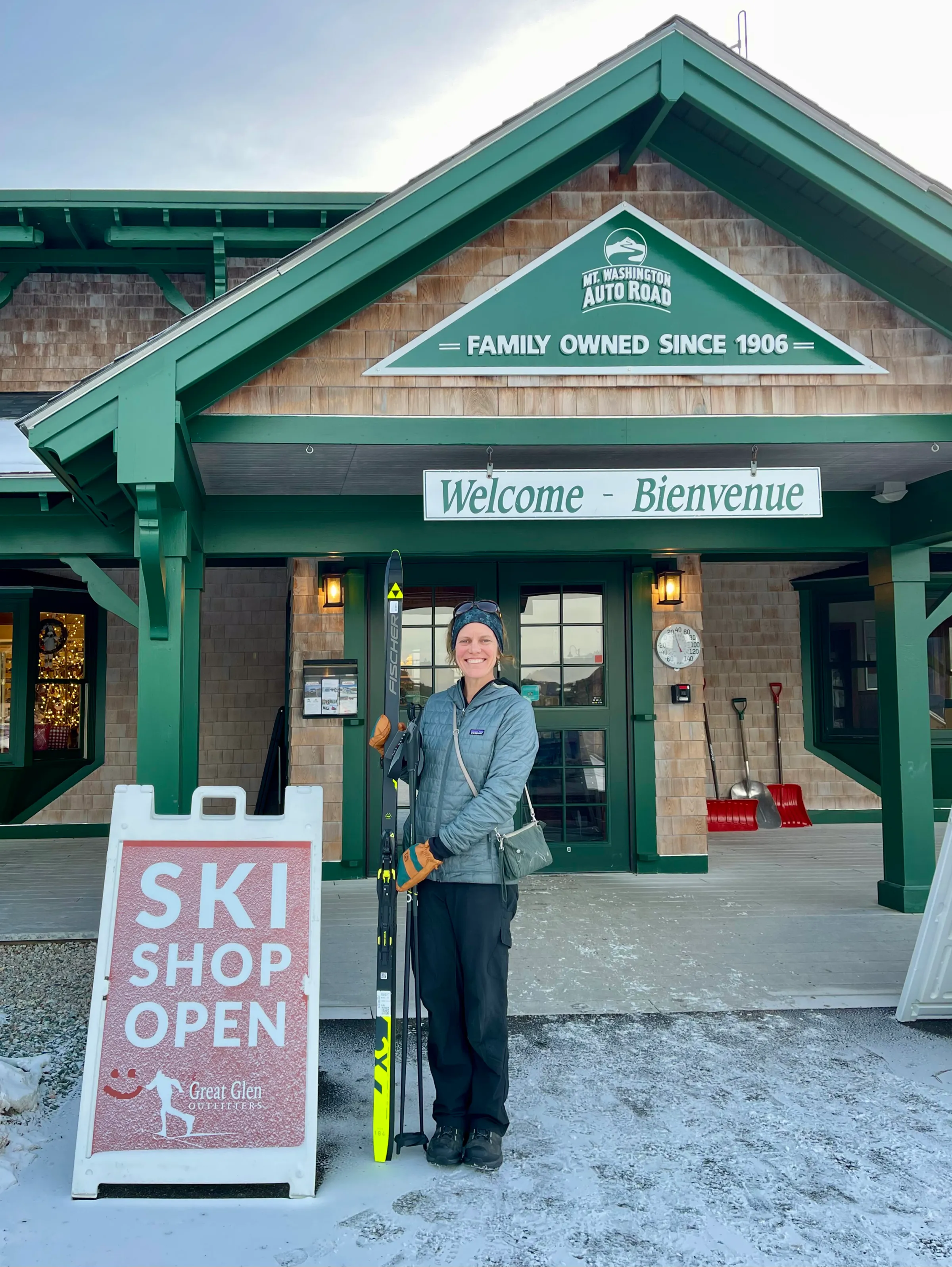 Person with skis stands by 'Ski Shop Open' sign at Mt. Washington Auto Road entrance.