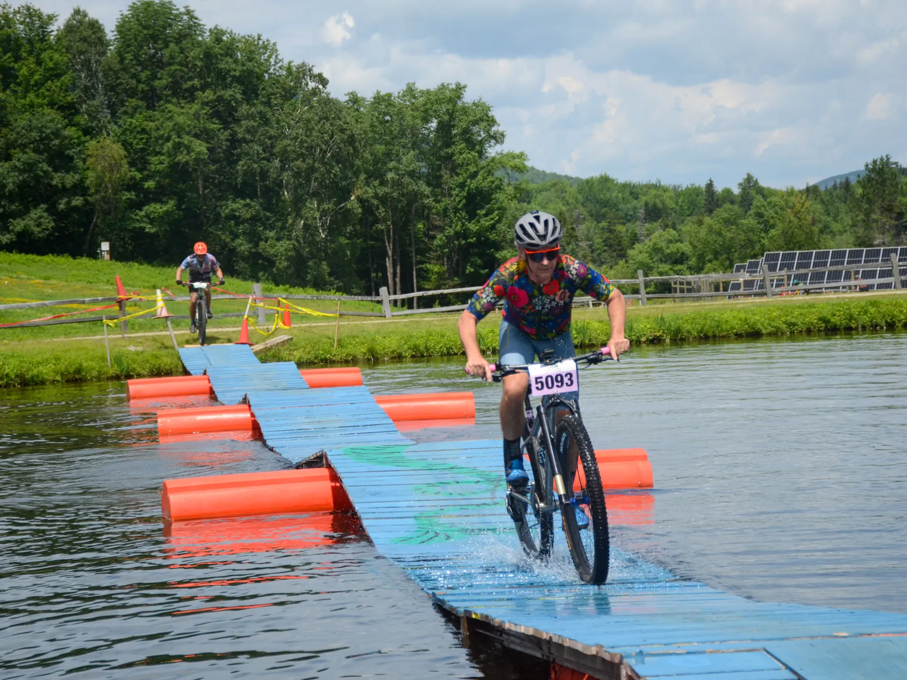 Cyclist rides over floating bridge on a lake during a race, with trees in the background.